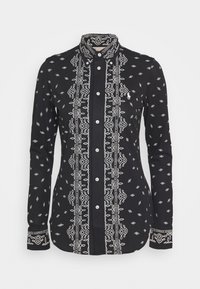 Svart skjorta med knappar som har vit geometrisk broderi längs framsidan och ärmsluten, med en strukturerad krage och långa ärmar.