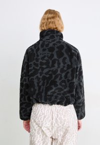 Fleece jakna z leopardskim vzorcem v črni in sivi barvi, z visokim ovratnikom in skrajšano zasnovo, ki se ponaša z teksturirano, mehko tkanino.