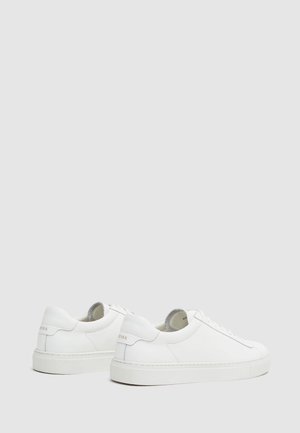 Par hvide lave lædersneakers med flade hvide såler, snørelukning foran og mærkede hæltabs på en ensartet lysegrå baggrund.