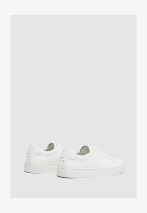 Par de zapatillas bajas blancas de cuero con suelas planas blancas, cordones en la parte delantera y lengüetas de talón con marca en un fondo liso gris claro.