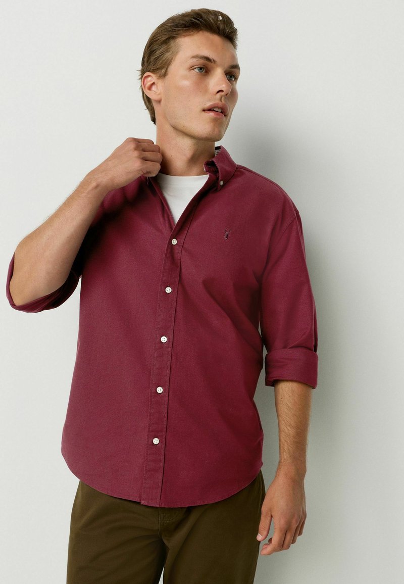 Camisa de botones color marrón con mangas arremangadas, hecha de un tejido suave, con un pequeño logo en el pecho y botones blancos. LLevada sobre una camiseta blanca.
