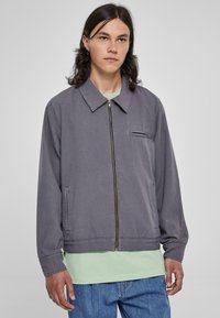 Chaqueta de algodón gris con cuello, cremallera frontal y un bolsillo en el pecho, llevada sobre una camiseta verde clara y combinada con jeans azules.
