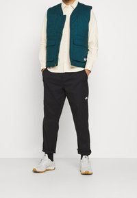 Turkos vadderad väst med två framfickor över en beige skjorta, tillsammans med svarta byxor och vita sneakers med ljusbruna sulor.