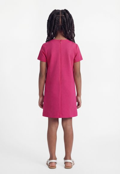 Παιδί με πλεξούδες, ντυμένο με ροζ φόρεμα κοντομάνικο μέχρι τα γόνατα και λευκά σανδάλια, στέκεται πλάτη σε απλό φόντο.