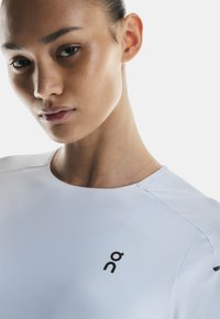 Haut de sport blanc avec une finition texturée, col rond et logo noir sur la poitrine gauche. Tissu lisse avec un design de couture ergonomique.