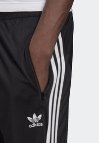 Pantaloni sportivi neri con tasca con zip, dotati di un design a tre strisce bianche lungo il lato e di un logo Adidas ricamato.