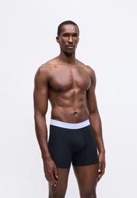 Fekete boxer alsónadrág puha, rugalmas derékkal, amelyen egy világoskék "Calvin Klein" logó látható. A modellen tónusos fizikum van.