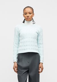 Femme aux cheveux tressés portant un pull en maille bleu clair à motifs sur un col roulé blanc et un pantalon plissé gris foncé, debout devant un fond blanc.