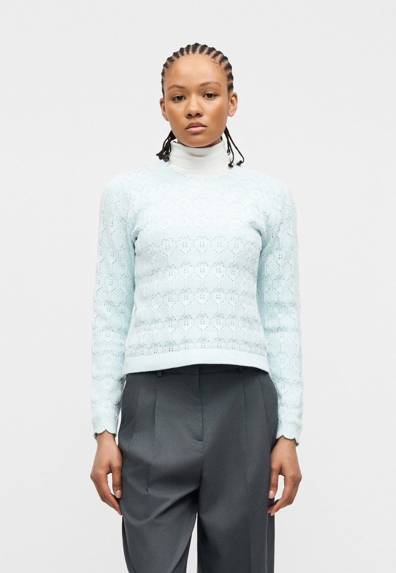 Femme aux cheveux tressés portant un pull en maille bleu clair à motifs sur un col roulé blanc et un pantalon plissé gris foncé, debout devant un fond blanc.
