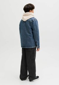 Jeans jakna u plavoj boji s opuštenim krojem, ispod koje se nalazi svijetlosiva dukserica s kapuljačom, crne traperice od kordura i crne patike.