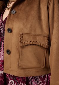 Veste en daim marron avec un design de poche cousue et deux boutons foncés. Texture douce, coupe raccourcie, portée avec une robe fleurie en dessous.