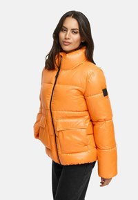 Een oranje pufferjack met een hoge kraag, voorzien van een ritssluiting aan de voorkant, twee grote zakken en een glanzende textuur. Zwarte accenten op de mouw.
