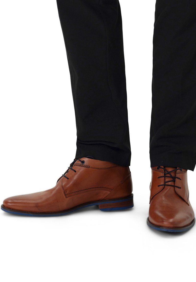 Bruine leren schoenen met een slank ontwerp, met een gladde afwerking, ronde neus en blauwe accenten op de zool. Draagt met zwarte broek.