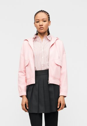 Young woman with braided hair wearing a pink zip-up jacket, striped button-up shirt, dark pleated skirt, and black pants, standing against white background.