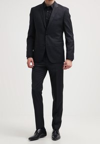 Uomo che indossa un completo nero su misura con una camicia nera abbinata e scarpe eleganti nere, in piedi contro uno sfondo bianco.