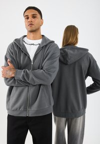 Felpa grigia con zip e cappuccio grande, dotata di una texture liscia e vestibilità comoda. Indossata sopra una maglietta bianca, abbinata a pantaloni scuri.