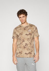 T-shirt en coton avec un motif camouflage dans des tons de beige et de marron, présentant un col rond et un petit logo blanc sur la poitrine.