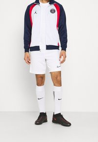 Witte sportjas met marineblauwe en rode accenten, voorzien van een logo. Gecombineerd met witte shorts, witte sokken met zwarte strepen en zwarte sportschoenen.
