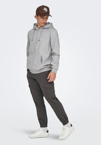 Sudadera gris con cremallera y bolsillos frontales, combinada con pantalones cargo oscuros que tienen bolsillos laterales y puños elásticos, usada con zapatillas blancas.