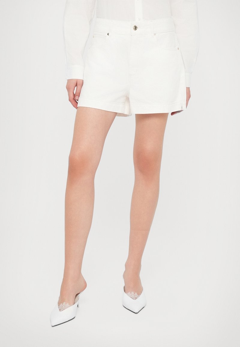 Femme portant un short blanc taille haute associé à un haut blanc à manches longues et des mules blanches à talons pointus avec un détail en dentelle.