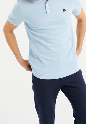 Mężczyzna w jasnoniebieskiej koszulce polo z krótkim rękawem z żółtym logo ptaka i ciemnoniebieskich spodniach, stojący z rękami za plecami.