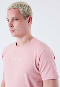 Camiseta corta de manga corta en color rosa, hecha de una tela suave. Presenta un logo en relieve "PXP" en el centro. Diseño ajustado con cuello redondo.