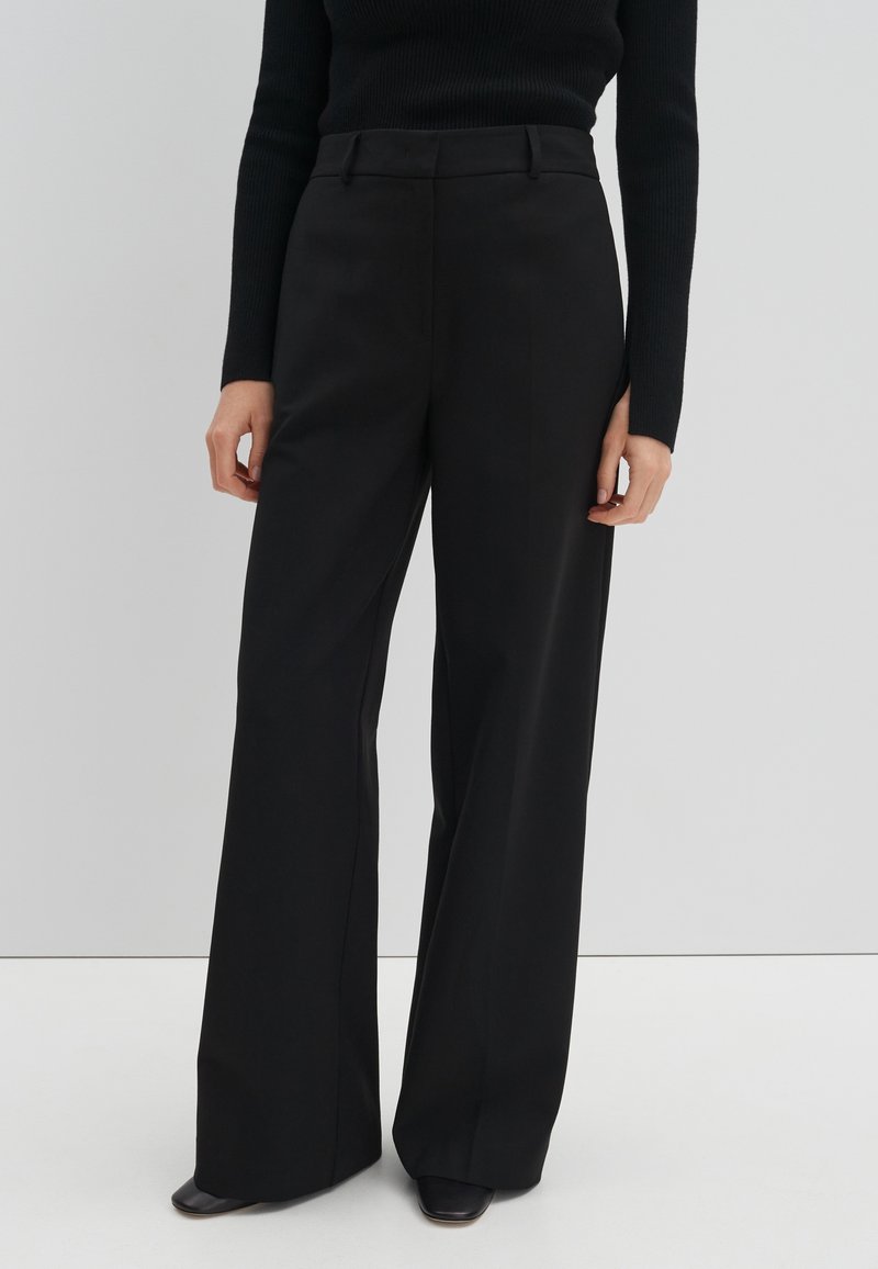 Personne portant un pantalon large taille haute noir et un haut noir ajusté à manches longues sur un fond clair uni.