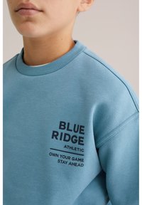 Oseba v svetlom modrem puloverju s črnim napisom "BLUE RIDGE ATHLETIC OWN YOUR GAME STAY AHEAD" na prsih.