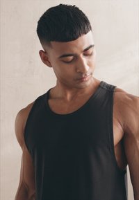 Fekete ujjatlan trikó textúrált felülettel és kerek nyakkivágással. A modellen izmos karok és enyhe oldalprofil látható.