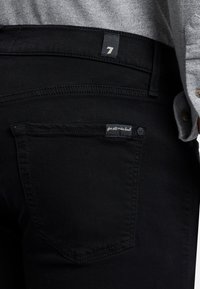 Sorte denim jeans med et nærbillede af baglommen, der har et lille mærke med "for all man kind" og en logo-knap.
