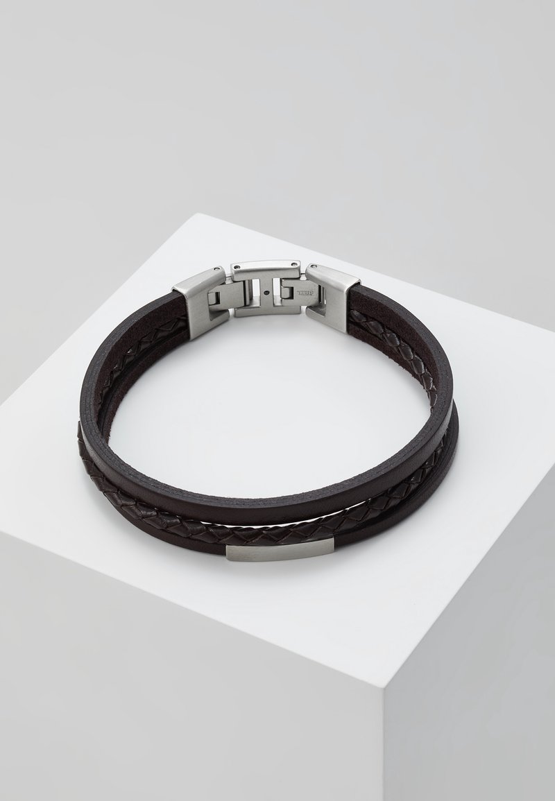 Bracelet double bande en cuir noir avec détail tressé, fermoir en métal argenté et plaque sur un socle blanc.