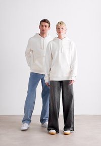 Deux modèles portant des sweat-shirts crème ; l'un associé à un jean bleu clair, l'autre avec un pantalon large gris foncé et des baskets blanches.
