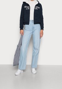 Felpa navy con zip e branding bianco, dolcevita bianco, jeans a gamba dritta azzurri chiari e sneakers bianche. Tiene un ombrello a righe.