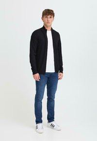 Svart zip-up kofta i textil, bärs över en vit t-shirt, tillsammans med blå jeans och vita sneakers med svarta ränder.