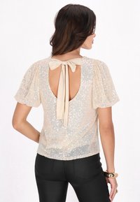 Kurzärmliges, beige Pailletten-Oberteil mit einem Rückensausschnitt und einer Schleifenverschluss. Verfügt über eine strukturierte Oberfläche und eine figurbetonte Passform.