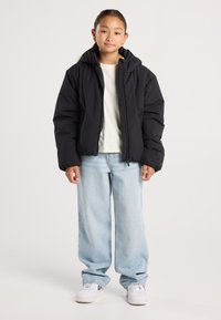 Young person standing wearing a black puffer jacket, cream shirt, light blue wide-leg jeans, and white sneakers on a plain background.