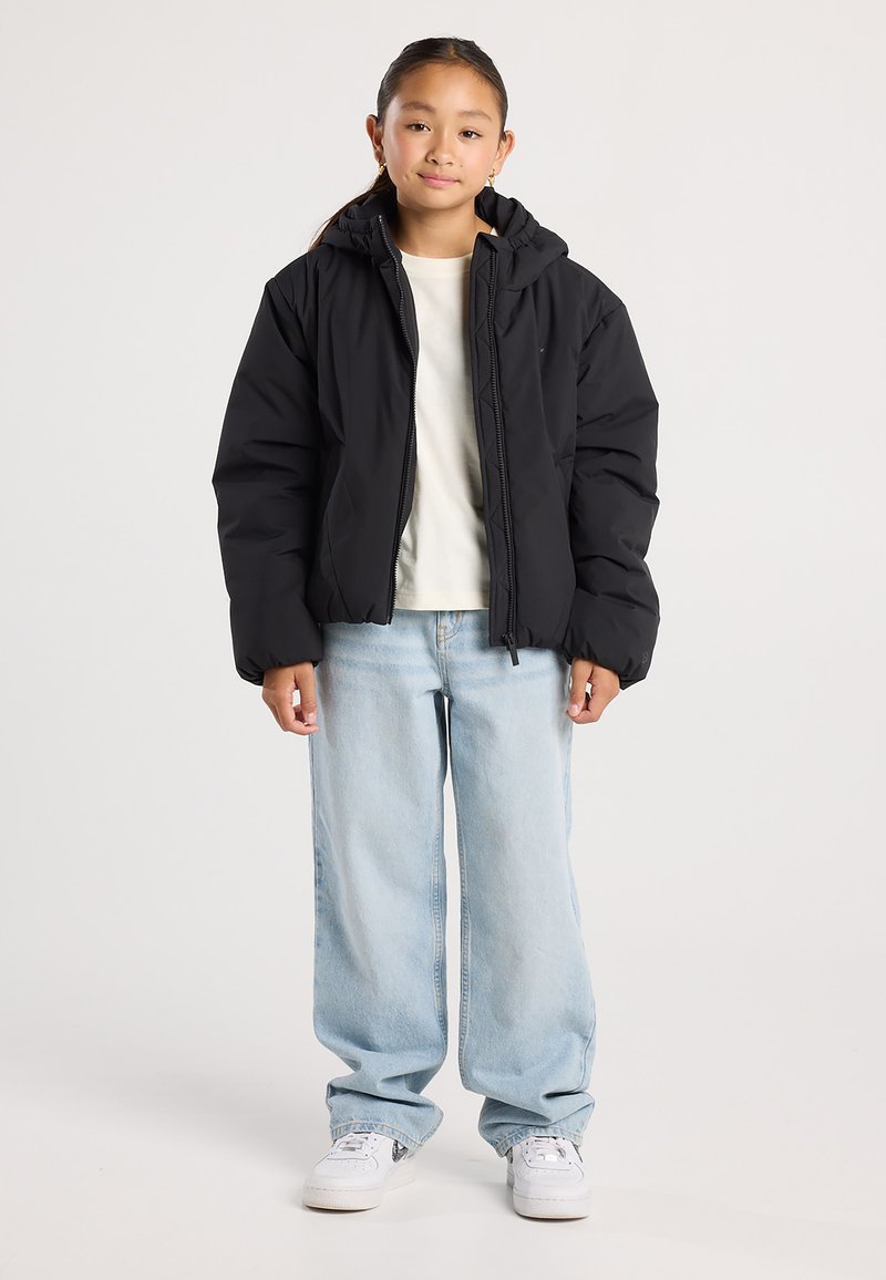 Young person standing wearing a black puffer jacket, cream shirt, light blue wide-leg jeans, and white sneakers on a plain background.