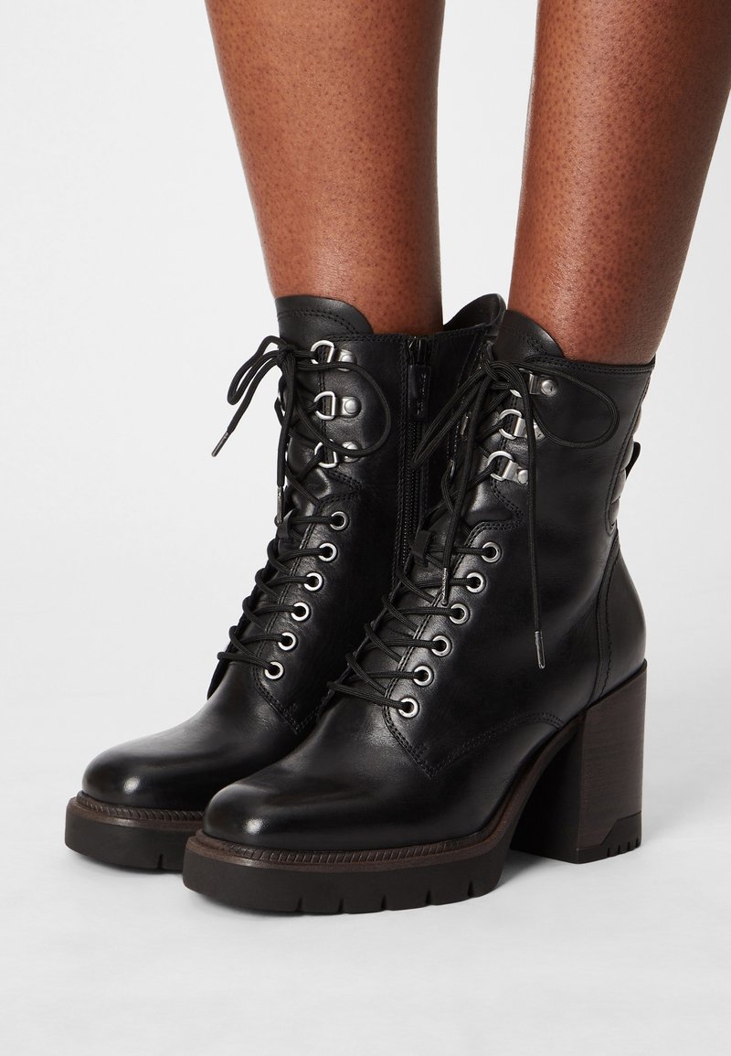 Bottines en cuir noir à lacets avec talons carrés épais portées sur des jambes à peau foncée sur un fond blanc.