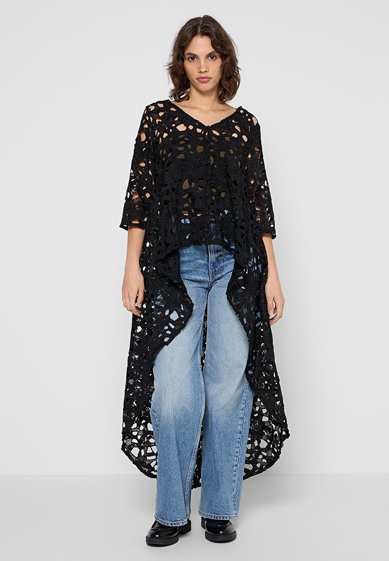 Frau, die ein schwarzes, asymmetrisches Spitzentop über einem braunen Unterhemd trägt, kombiniert mit hellblauen, weiten Jeans und schwarzen Schuhen.