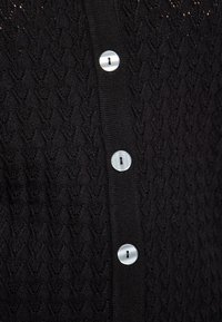 Černý pletený cardigan s texturovaným vzorem ve tvaru V, se řadou kulatých stříbrných knoflíků na přední straně.