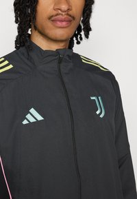 Veste noire zippée avec un col haut, présentant trois rayures jaunes sur les épaules et des logos bleu clair sur la poitrine.