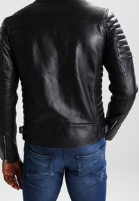 Svart lädermotorcykeljacka med kviltade axelpartier, blixtlås vid ärmsluten och en skräddarsydd passform. Bärs över blå jeans.