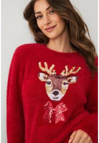Rode, pluizige trui met een rendiergezicht ontwerp, voorzien van pailletten details en een rode strik. Zachte textuur en losvallende pasvorm.
