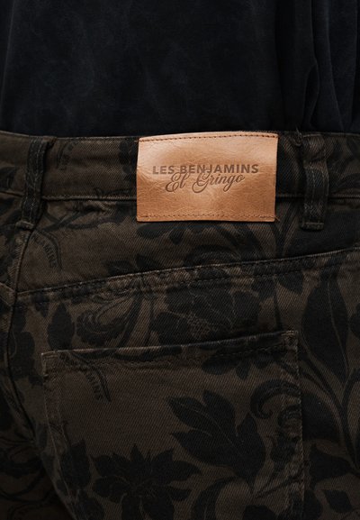 Καφέ δερμάτινο μπατς με ετικέτα «LES BENJAMINS El Gringo» σε τσέπη σκούρου παντελονιού με λουλουδάτο μοτίβο, κάτω από μαύρο πουκάμισο.