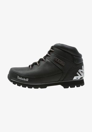 Stivali da trekking neri con tomaia in pelle testurizzata, suole in gomma e occhielli in metallo. Design geometrico bianco ben visibile sul tallone. Logo Timberland in evidenza.