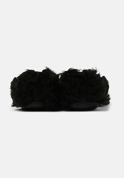 Un paio di scarpe slip-on nere, con texture pelosa e suole in gomma, viste da dietro su uno sfondo grigio semplice.