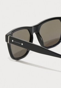 Óculos de sol pretos com armações retangulares, lentes de tom escuro e acabamentos prateados nas hastes. Apresenta logo na lateral. Textura suave.