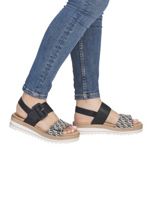 Person wearing blue jeans and black-and-white woven sandals with ankle straps and white rubber soles.