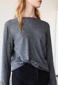 Szary top z długim rękawem o luźnym kroju i subtelnej fakturze; posiada okrągły dekolt i supełkowy detal z przodu, zestawiony z czarnym dołem.