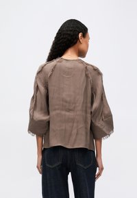 Frau mit langen lockigen Haaren, die eine locker sitzende braune Bluse mit ausgefransten Kanten und dunkelblaue Jeans trägt, mit dem Rücken zu einer einfarbigen Hintergrund.